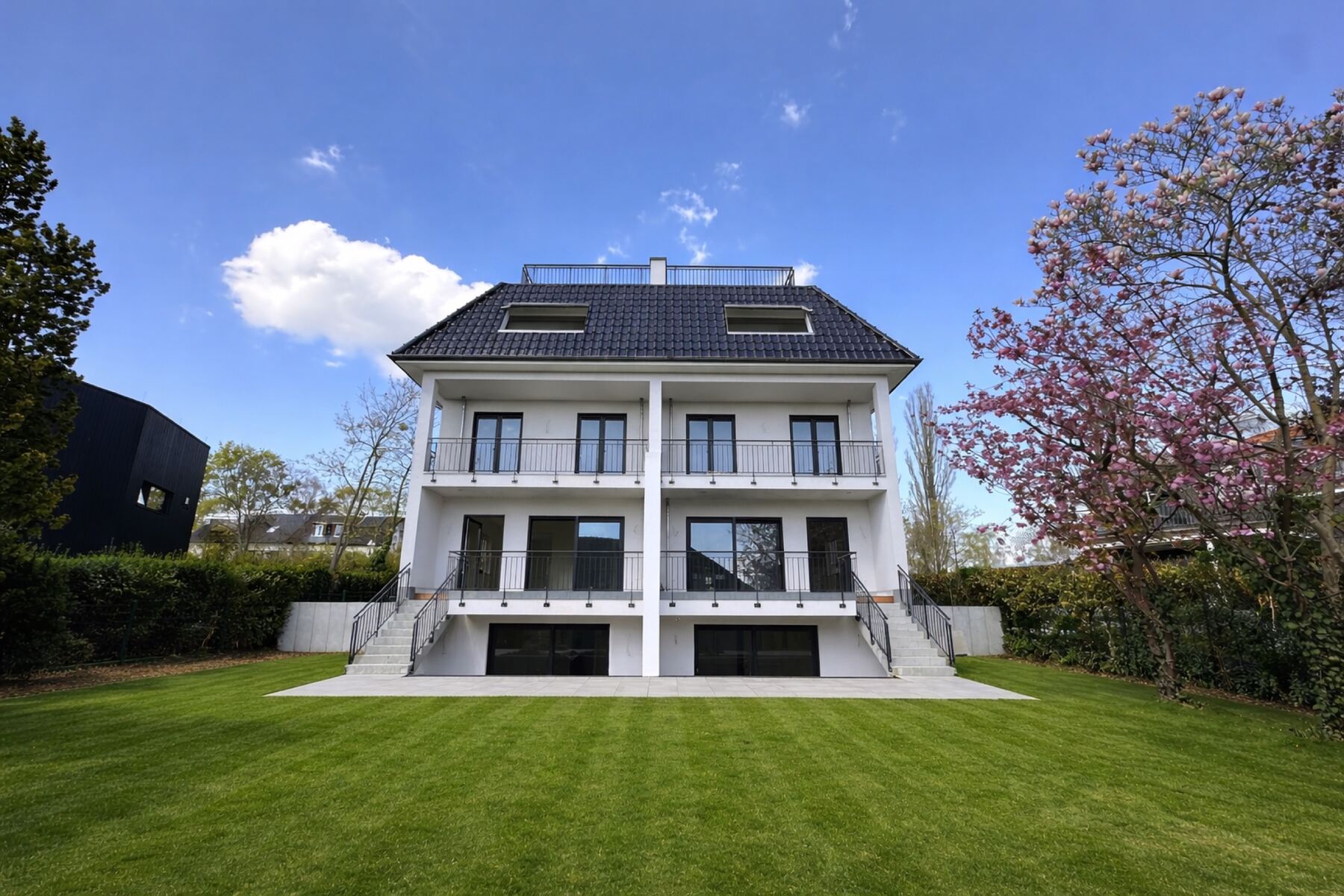 Weiße Doppelhaushälfte mit Balkonen und Dachterrasse, davor ein gepflegter Rasen und eine blühende Magnolie